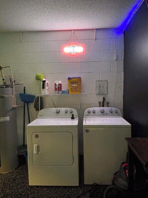 Ein Waschraum mit zwei Waschmaschinen mit Frontlader, einem Regal mit Reinigungsmitteln und einem roten Neonlichtschild an der Wand.