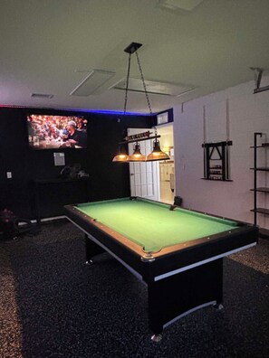 Ein Billardtisch mit grüner Spieloberfläche, eine Hängelampe und im Hintergrund ein Fernseher, der ein Sportevent zeigt.