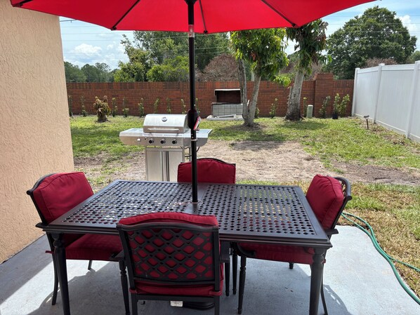 Eine Außenpatio mit Tisch und Stühlen, einem Grill und einem roten Sonnenschirm.