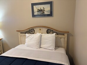 Ein Bett mit weißer Bettwäsche und zwei Kissen, ein gerahmtes Bild darüber sowie ein hölzernes Kopfende mit geschwungenen Verzierungen.