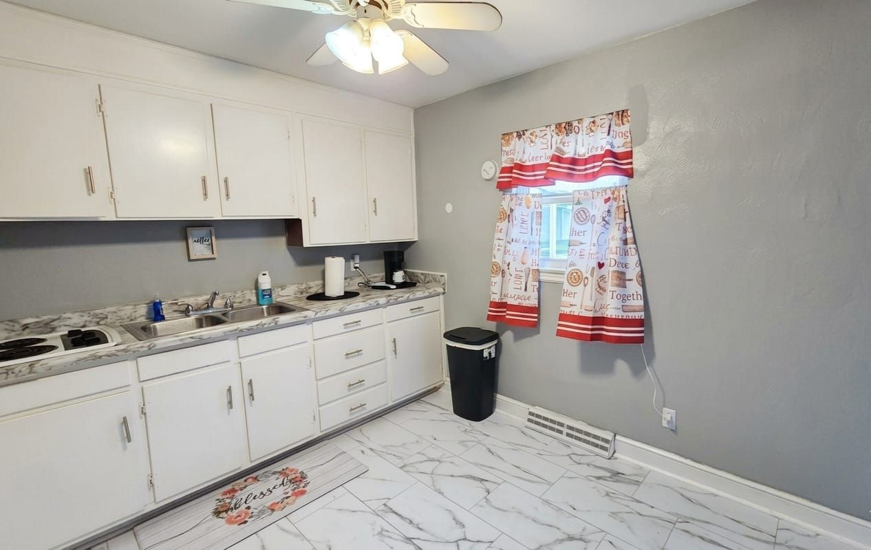 A kitchen with white cabinets, a marble countertop, a ceiling fan, a window with a patterned curtain, and a trash can.