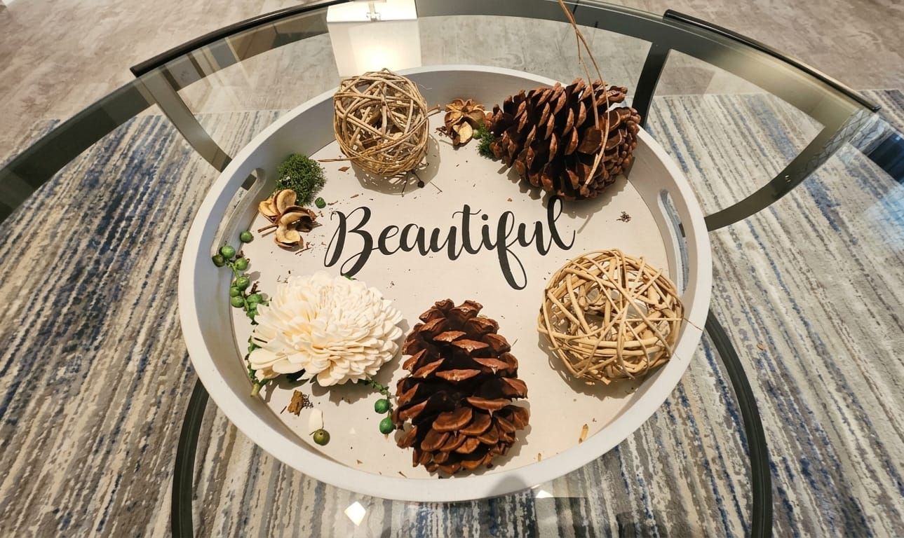 A round table with a glass top, decorated with pinecones, a woven ball, and white flowers. The word 'Beautiful' is written on the table.