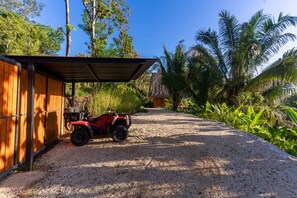 Ein Schotterweg führt zu einem Holzhaus mit Metallbedachung, umgeben von tropischer Vegetation.