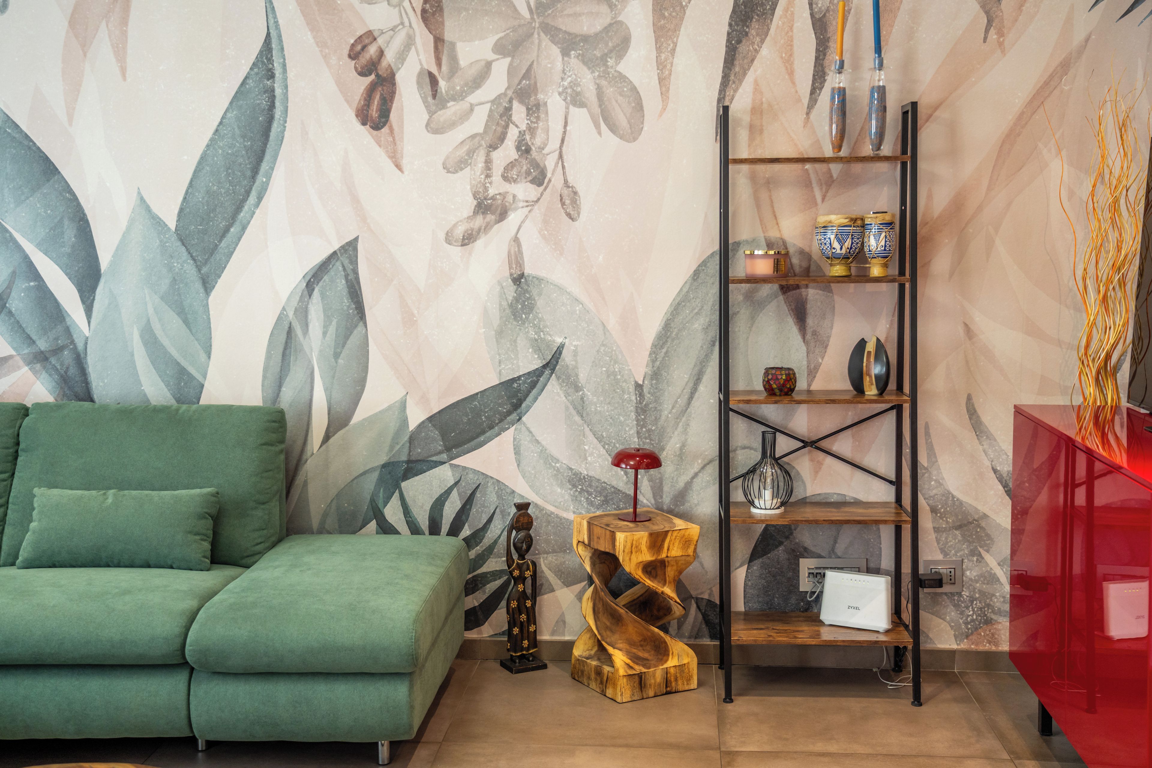 Una habitación con un sofá verde, una mesita de madera con una lámpara roja, una estantería espiral de madera con adornos y una pared con un diseño de hojas.