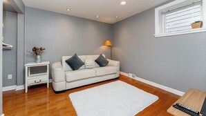 A modern living room with a white sofa, a small side table, a lamp, and a window with a plant. - Garden suite by DSOUZA (Vancouver)
