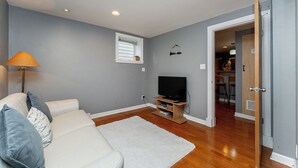 A living room with a sofa, a television on a stand, a white rug, and a door leading to another room. - Garden suite by DSOUZA (Vancouver)