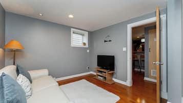 A living room with a sofa, a television on a stand, a white rug, and a door leading to another room.