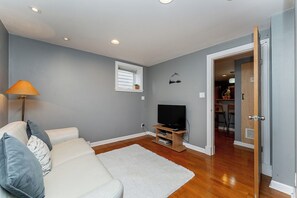 A living room with a sofa, a television on a stand, a white rug, and a door leading to another room. - Garden suite by DSOUZA (Vancouver)