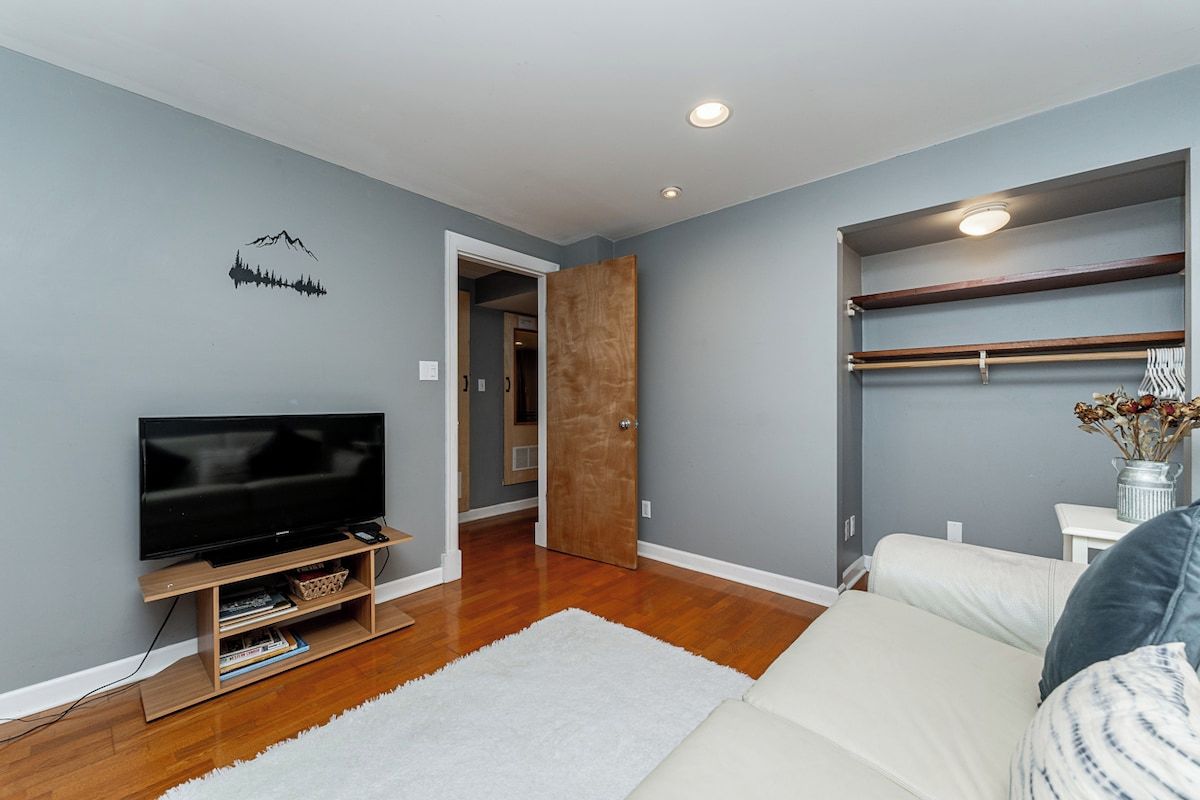 A modern living room with a flat-screen TV, wooden flooring, a white rug, and a built-in shelving unit.
