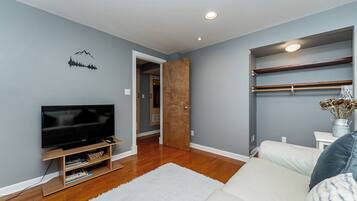 A modern living room with a flat-screen TV, wooden flooring, a white rug, and a built-in shelving unit.