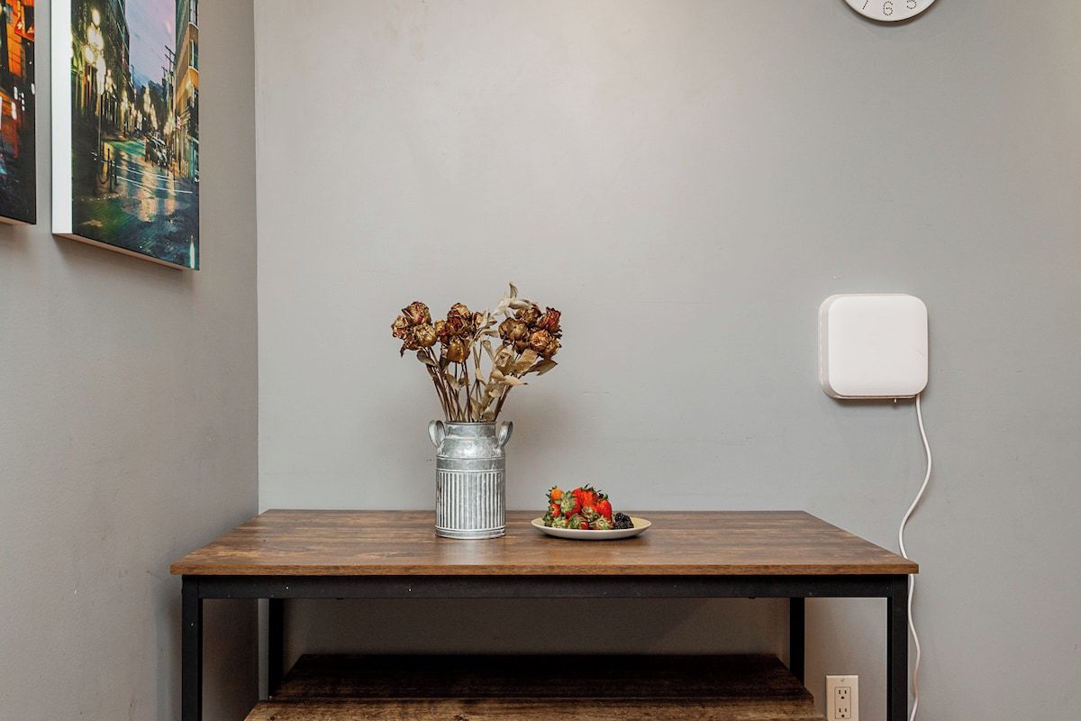 Ein Tisch mit einer Vase getrockneter Blumen, einer kleinen Teller mit Essen, einer Steckdose und einem Bild an der Wand.