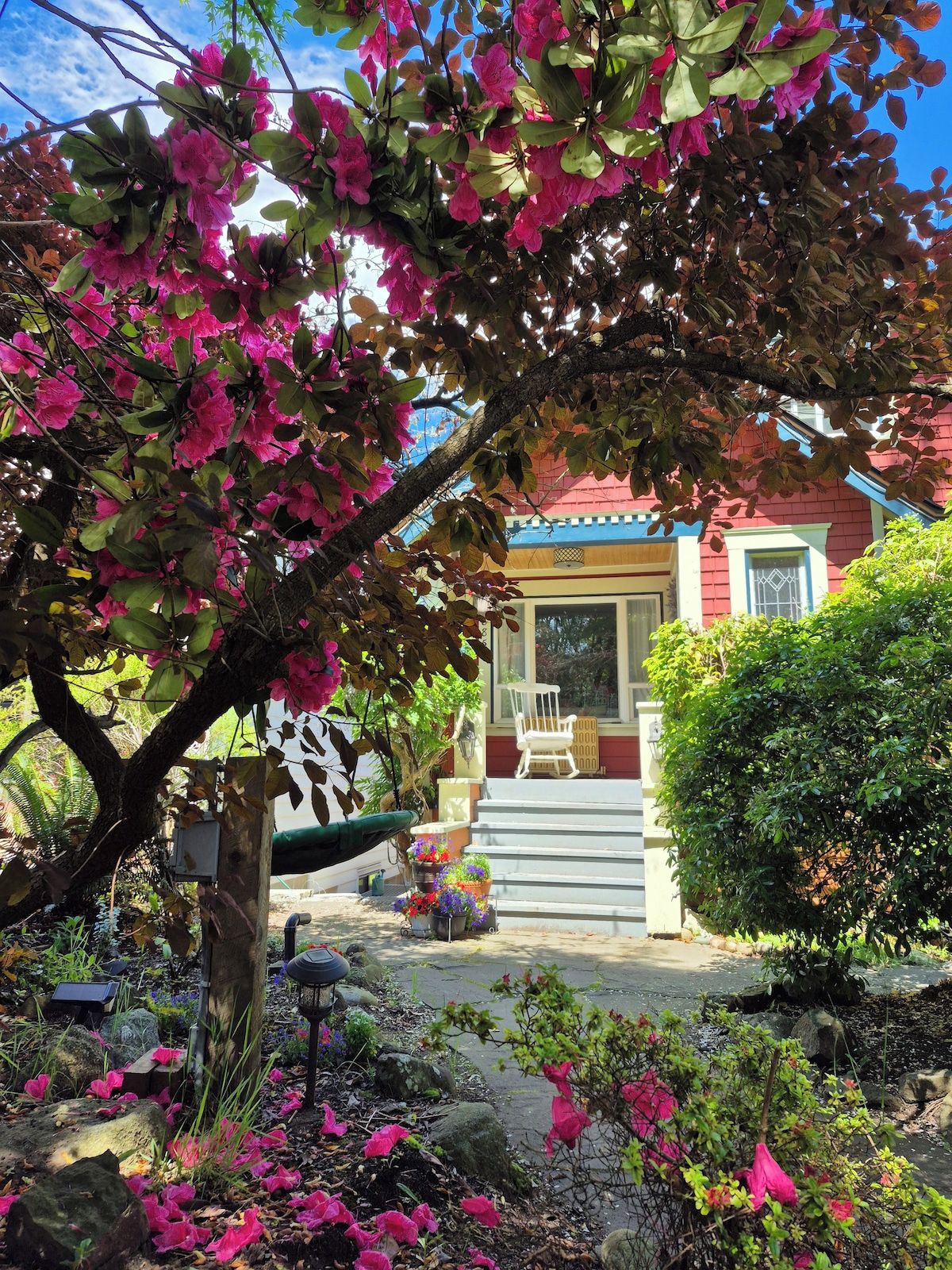 Ein Haus mit Veranda, einem Stuhl und einem Garten mit rosa Blumen.