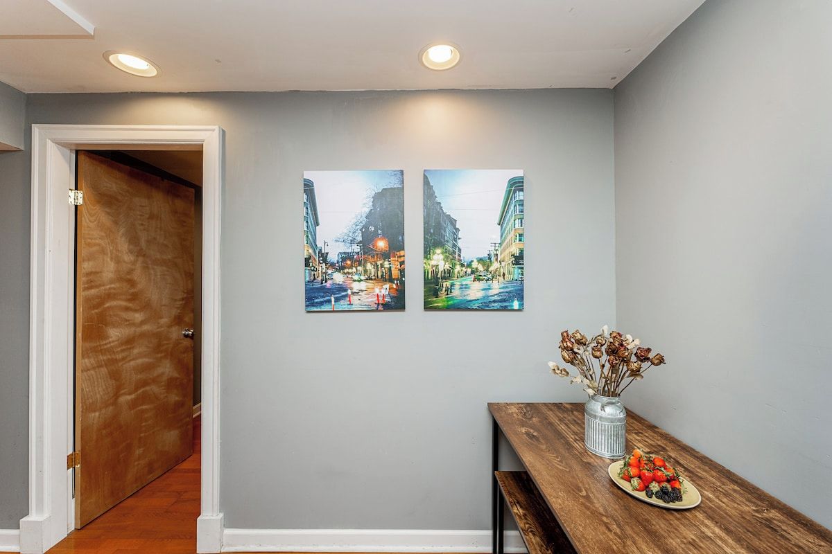 A hallway with a wooden door, a dining table with a plate of fruit, and two cityscape paintings on the wall.
