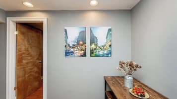 A hallway with a wooden door, a dining table with a plate of fruit, and two cityscape paintings on the wall.