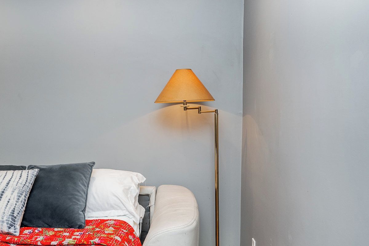 A bed with a red patterned bedspread, a white pillow, and a grey pillow. A floor lamp with a beige lampshade is positioned to the right of the bed.