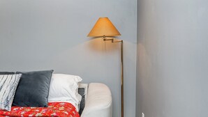 A bed with a red patterned bedspread, a white pillow, and a grey pillow. A floor lamp with a beige lampshade is positioned to the right of the bed. - Garden suite by DSOUZA (Vancouver)