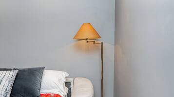 A bed with a red patterned bedspread, a white pillow, and a grey pillow. A floor lamp with a beige lampshade is positioned to the right of the bed.