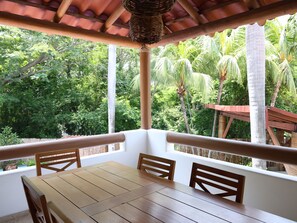 Een houten tafel en stoelen onder een pergola met uitzicht op weelderige begroeiing.