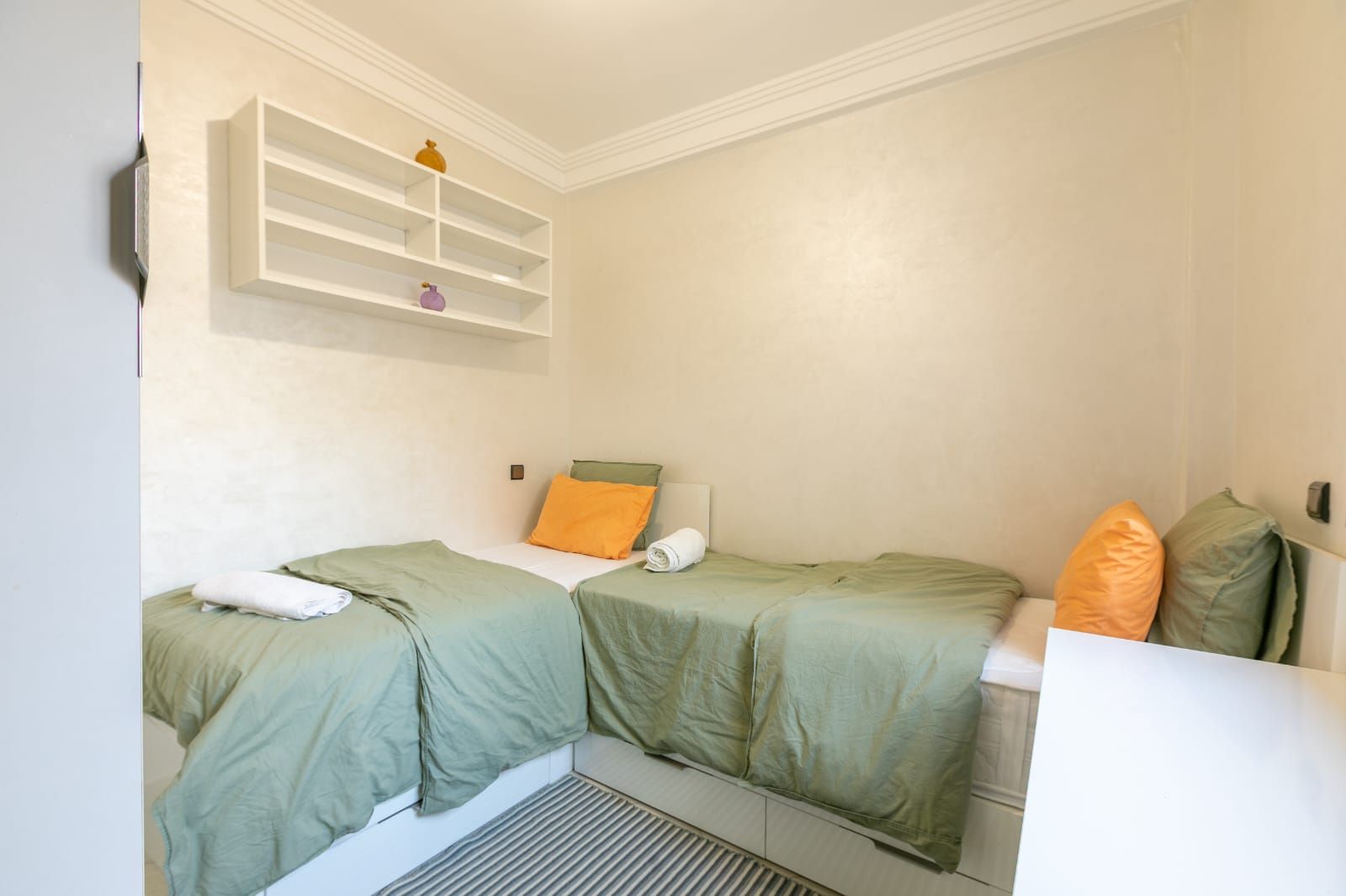 A room with two beds, a shelf with decorative items, and a white wall.