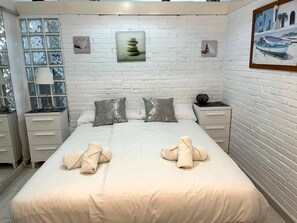 A neatly made bed with two pillows and two towels, a white brick wall, and a painting of boats. - Studio in Nerja Near Carabeillo Beach (Nerja)