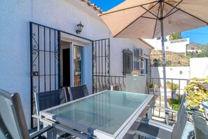 An outdoor patio with a glass table and chairs, a beige umbrella, and a view of a hillside. - Holiday Home in Nerja With Sea Views (Nerja)