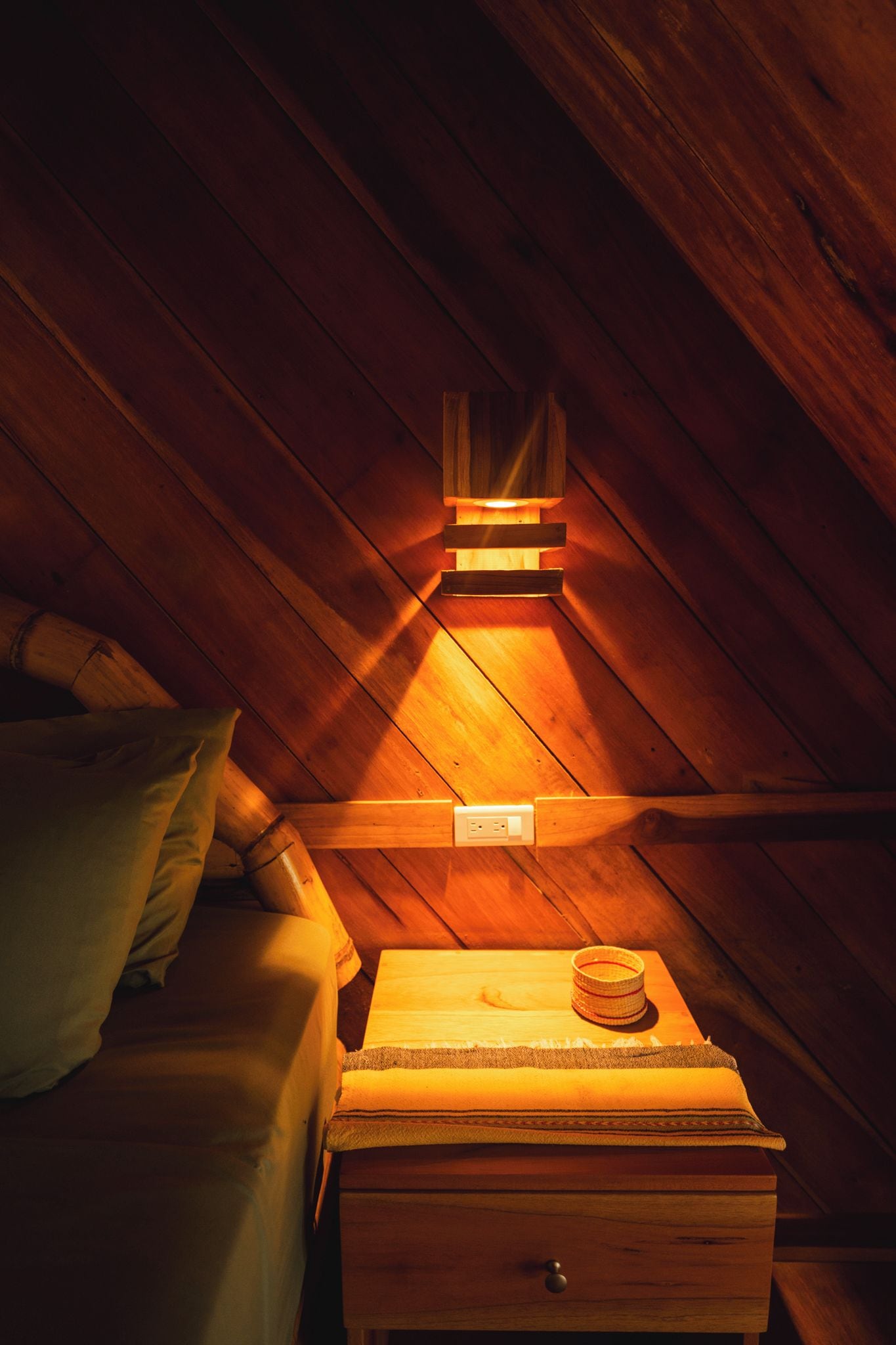 Una cama litera de madera con cabecera, una mesita de noche con lámpara y un pequeño cofre de madera.