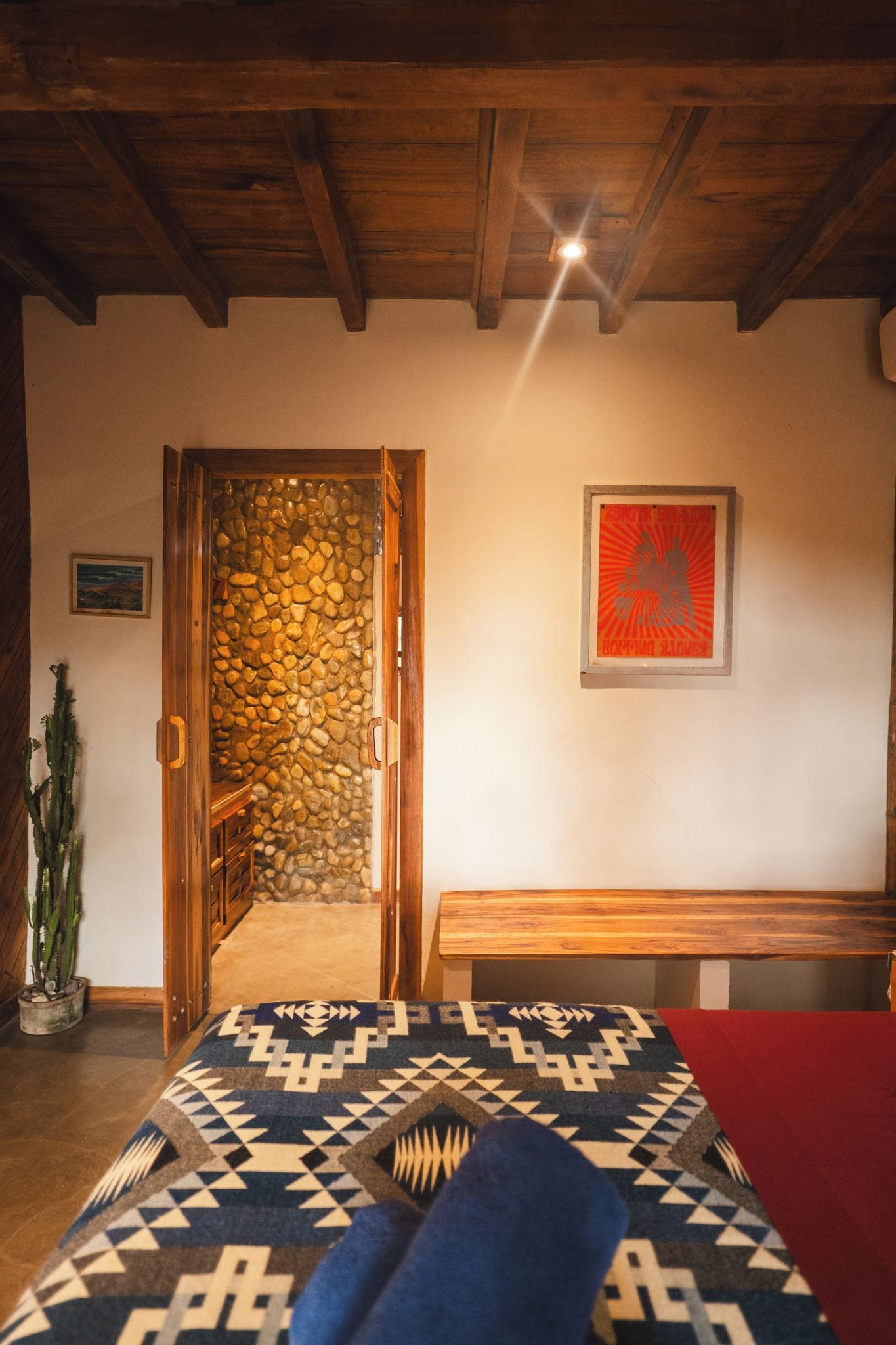 Una habitación con techo de madera, pared de piedra, puerta de madera, un cuadro enmarcado, un banco y un tapete estampado.