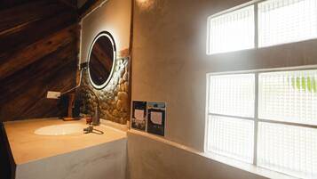 A bathroom with a stone wall, a round mirror, a sink, and a window with blinds.
