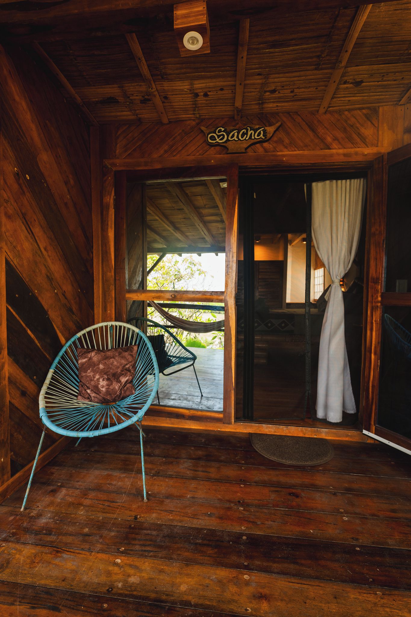 Interior de una cabaña de madera con una silla hamaca, un ventanal corredizo y un letrero que dice 'Sacha'.
