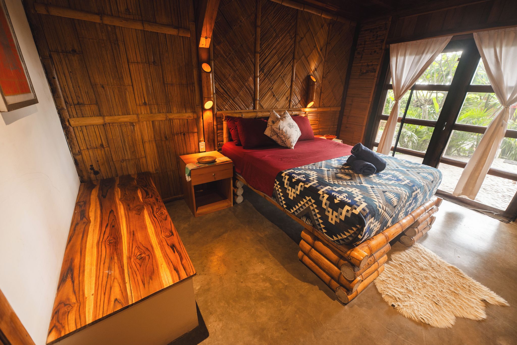 A room with a bed, a nightstand, a wooden bench, and a window with curtains.