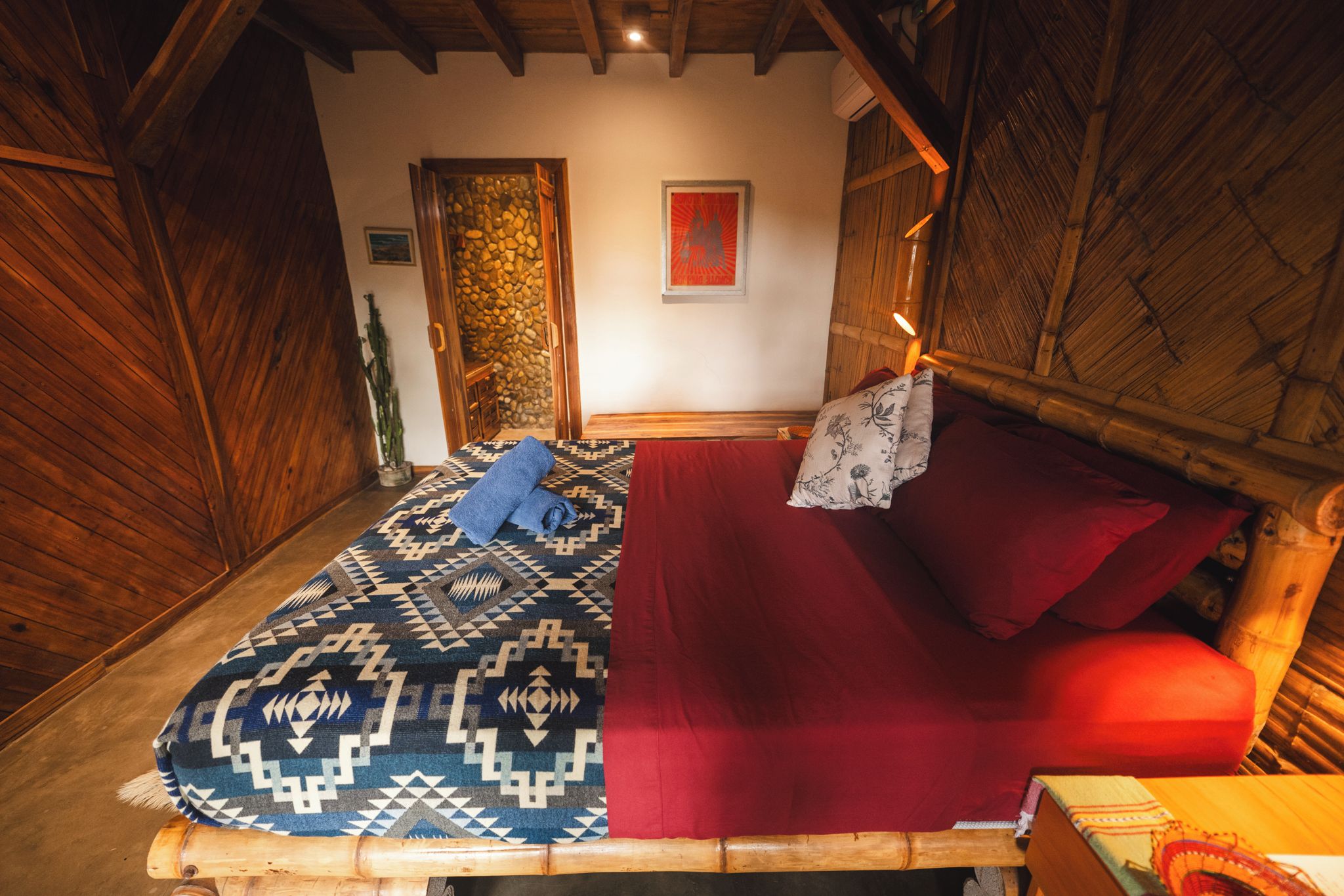 A wooden bed with a patterned blanket and red pillows in a room with a stone wall and wooden ceiling beams.