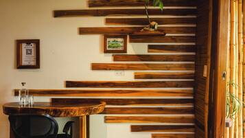 A room with a slatted wooden wall, a desk with a chair, a small bed, and a door leading outside.