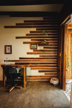 A room with a slatted wooden wall, a desk with a chair, a small bed, and a door leading outside.