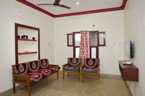 A living room with a sofa, two armchairs, a ceiling fan, a television, and a shelf with decorative items.