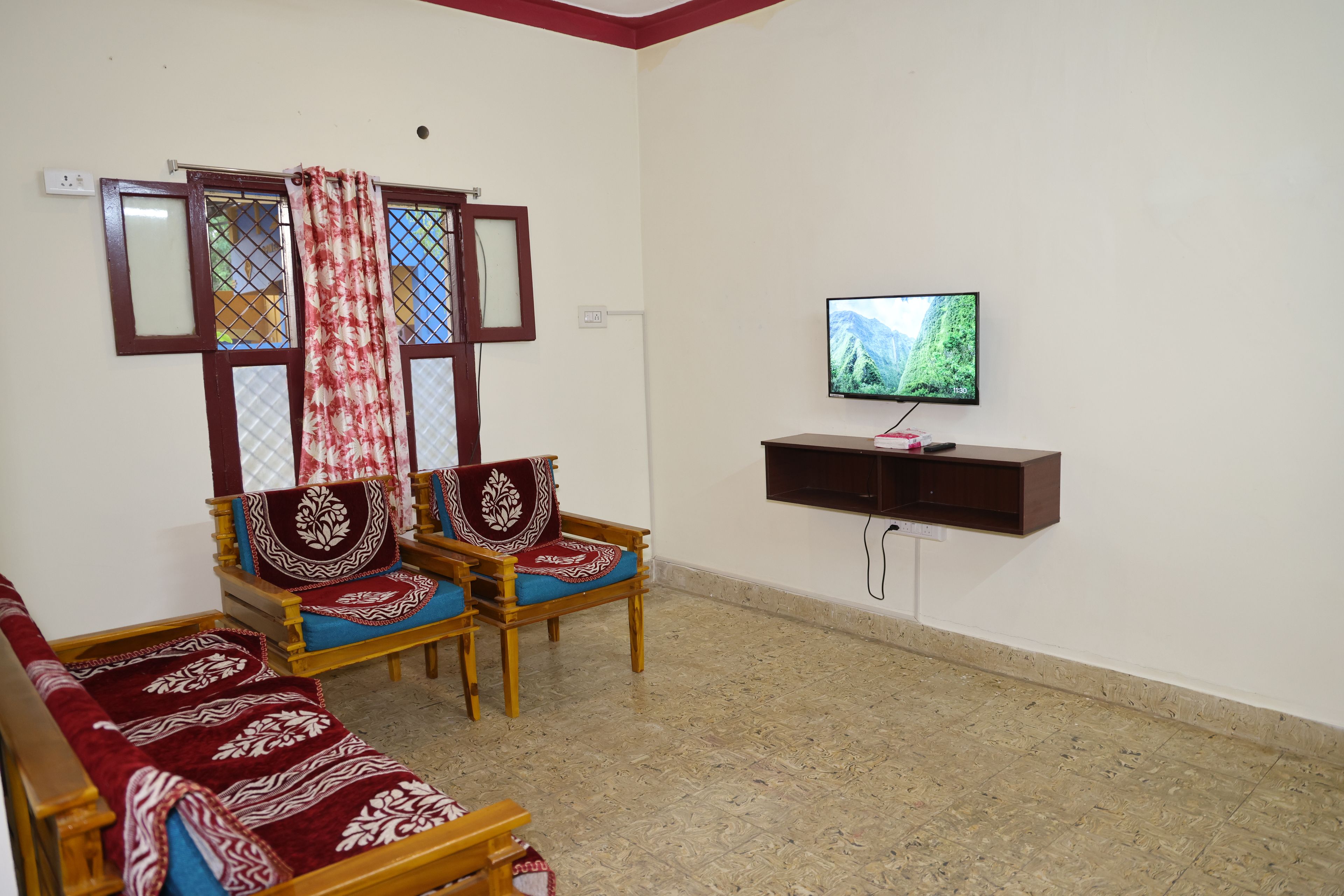 A living room with two wooden sofas, a television on a wall-mounted stand, and a window with a curtain.