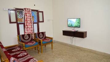 A living room with two wooden sofas, a television on a wall-mounted stand, and a window with a curtain.