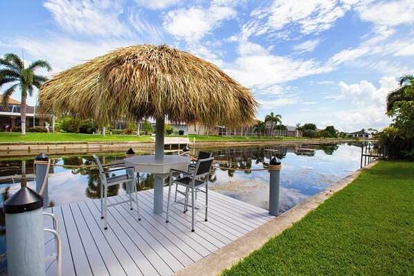 A deck with a thatched umbrella, a table, and chairs by a calm water body, surrounded by greenery and buildings. - Happy Oasis- Relax, Swim & Unwind in Your Tropical Cape Coral Retreat (Cape Coral)