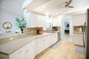 A modern kitchen with white cabinets, a large island, a ceiling fan, and a view to the living area. - Happy Oasis- Relax, Swim & Unwind in Your Tropical Cape Coral Retreat (Cape Coral)