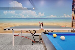 Taula de ping-pong amb un mural de fons de platja, taula de billar amb pilotes i un cable vermell.