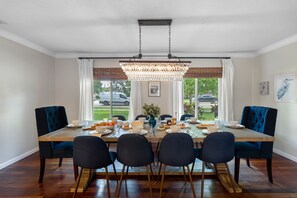 Una sala menjador amb taula de fusta preparada per a sis persones, envoltada de cadires tapissades fosques. Una gran lluminària penjant del sostre, i l'espai té grans finestres amb cortines blanques.