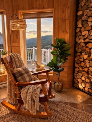 A wooden rocking chair with a cushion, a potted plant, a table with a candle and glasses, and a view of the outdoors through large windows. - Comfort & Nature - Alix (Saint-David-de-Falardeau)