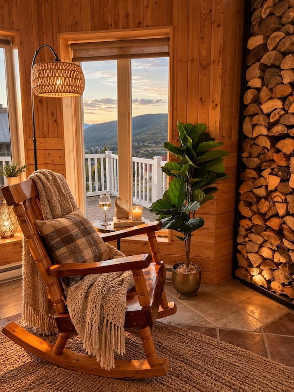 A wooden rocking chair with a cushion, a potted plant, a table with a candle and glasses, and a view of the outdoors through large windows.