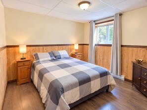 A bedroom with a checkered bedspread, wooden paneling, a nightstand with a lamp, a dresser, and a window with curtains. - Comfort & Nature - Alix (Saint-David-de-Falardeau)