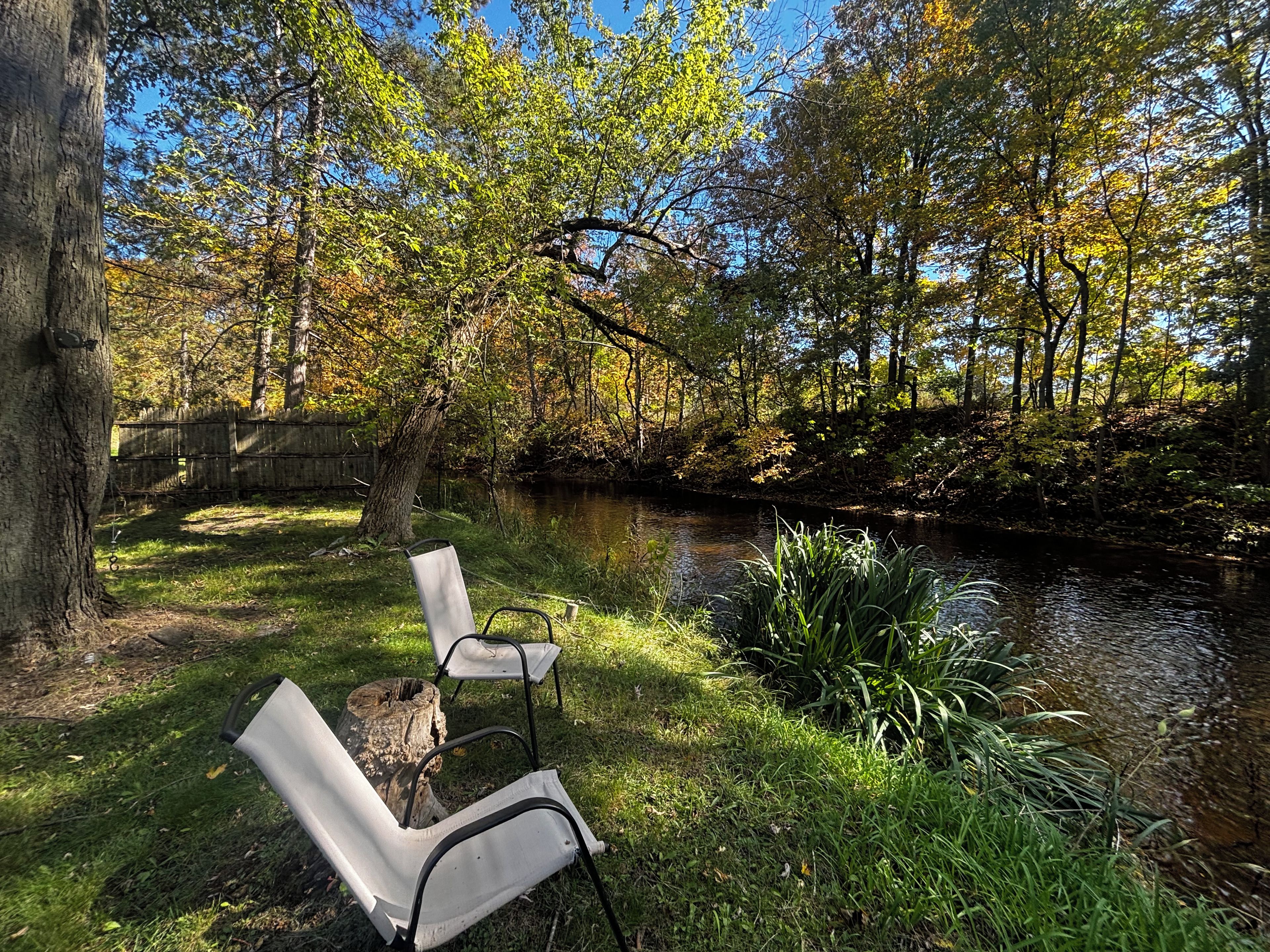 Een rustgevende omgeving buiten met een rivier, twee witte stoelen en weelderige begroeiing.