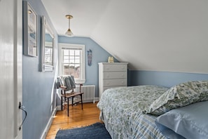 A bedroom with a sloped ceiling, a radiator, a wooden chair, a white dresser, and a bed with patterned bedding.