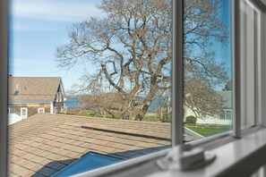 A view through a window of a residential area with a large tree, a tiled roof, and houses in the background. - Historic home on Bailey Island with ocean views. Steps from the Giant's Stairs. (Bailey Island)