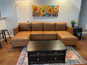 A sectional sofa with a matching ottoman, a dark wooden dresser, a colorful rug, and a painting of sunflowers on the wall. - Home SWEET Home away from home! Selma, TX! (Selma)