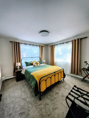 Ein Schlafzimmer mit einem Bett, einem Nachttisch, einer Lampe, einem Stuhl und einem Fenster mit Vorhängen.