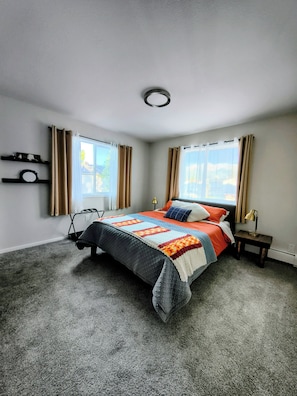 A bedroom with a bed, two bedside tables, a window with curtains, and a wall-mounted shelf.