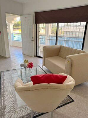 Sala de estar com sofá bege, mesa de centro de vidro com vaso e almofada vermelha, e vista para o exterior através de uma grande janela com grade de metal.